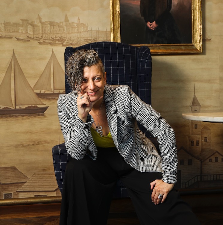 Emy Tafelski in a houndstooth blazer, chin on hand, perched in a plaid wingback chair against a nautical mural